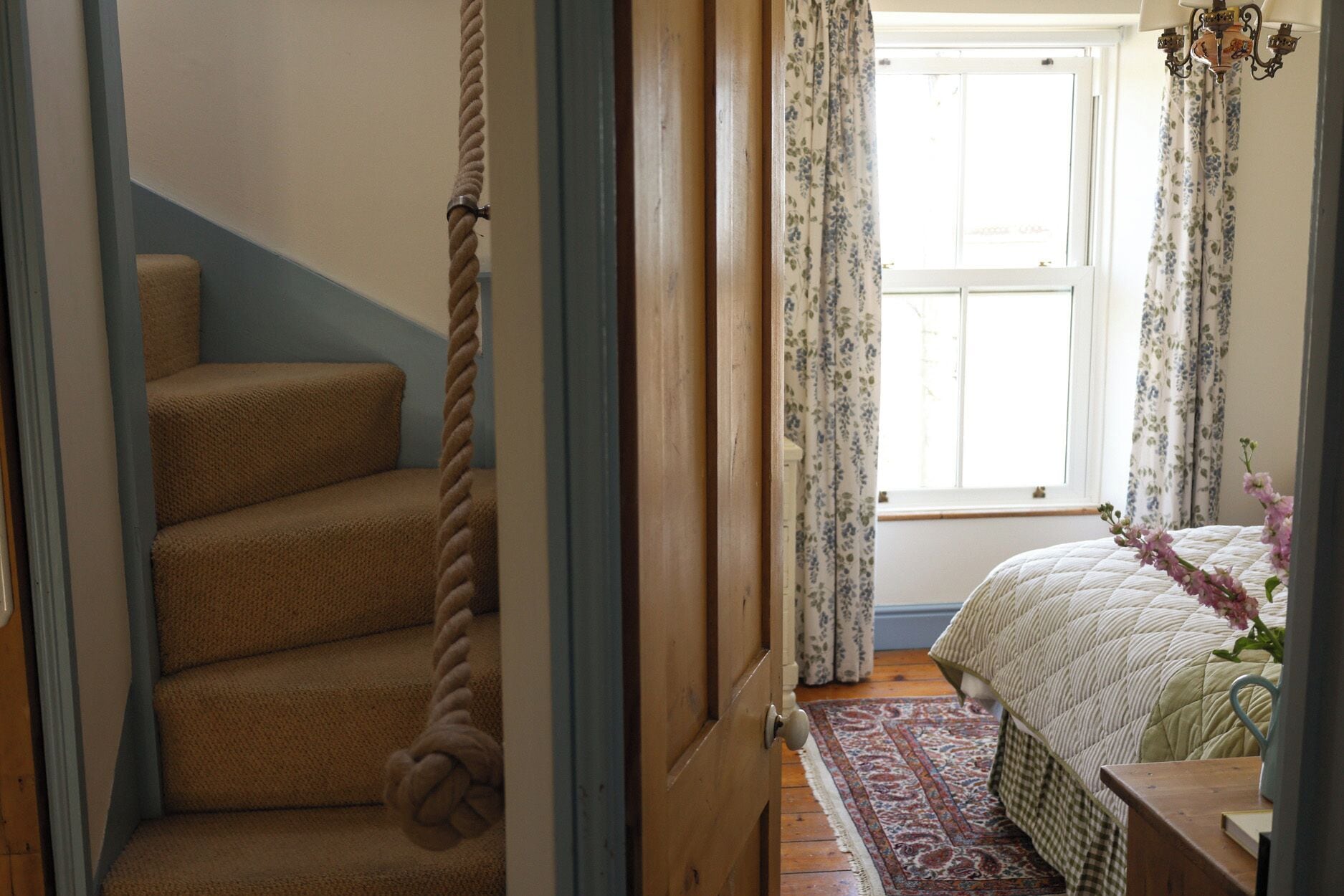 Un dormitorio con cama, ventana con cortinas florales, escalera de madera con cuerda y alfombra estampada.