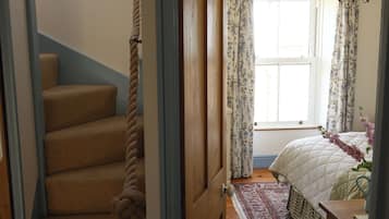 Un dormitorio con cama, ventana con cortinas florales, escalera de madera con cuerda y alfombra estampada.