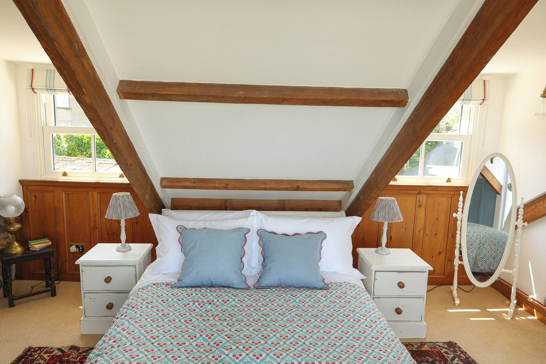 Un dormitorio con techo inclinado, vigas de madera, cama con ropa de cama estampada, dos mesitas de noche con lámparas, un espejo y una ventana con vistas.