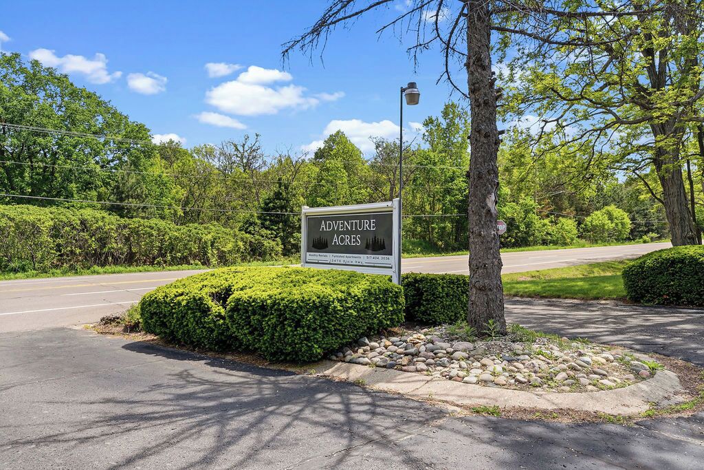 A sign for 'Adventure Acres' with a landscaped area featuring a circular stone pattern, a tree, and a street in the background.