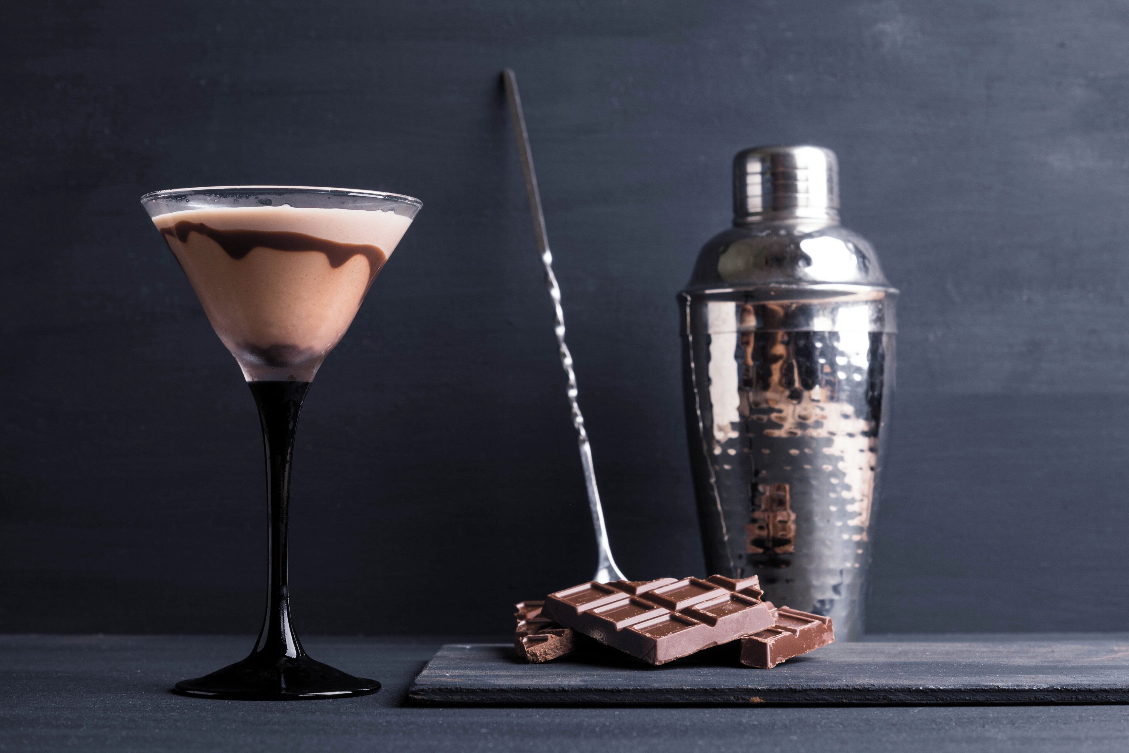 Una copa de martini con una bebida de chocolate, una barra de chocolate y un coctelero sobre una superficie oscura.