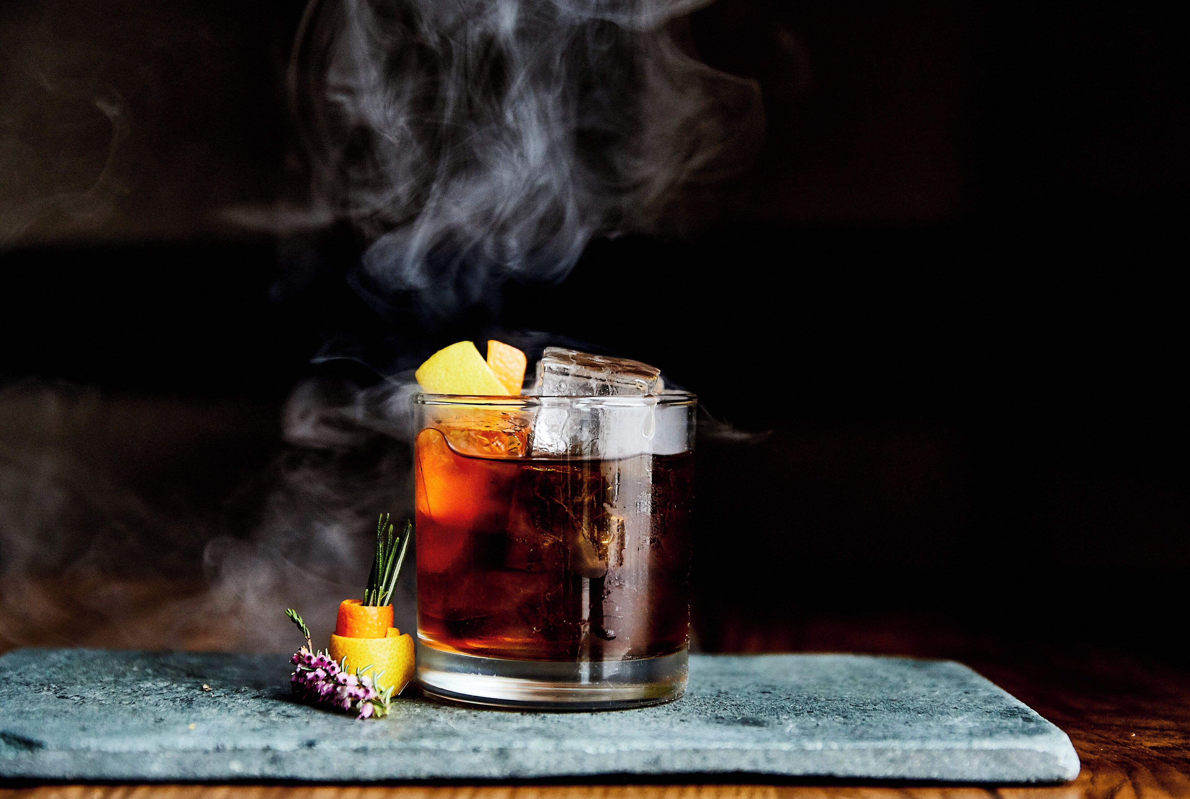 A glass of amber liquid with ice cubes and a lemon slice, surrounded by smoke, on a slate surface.