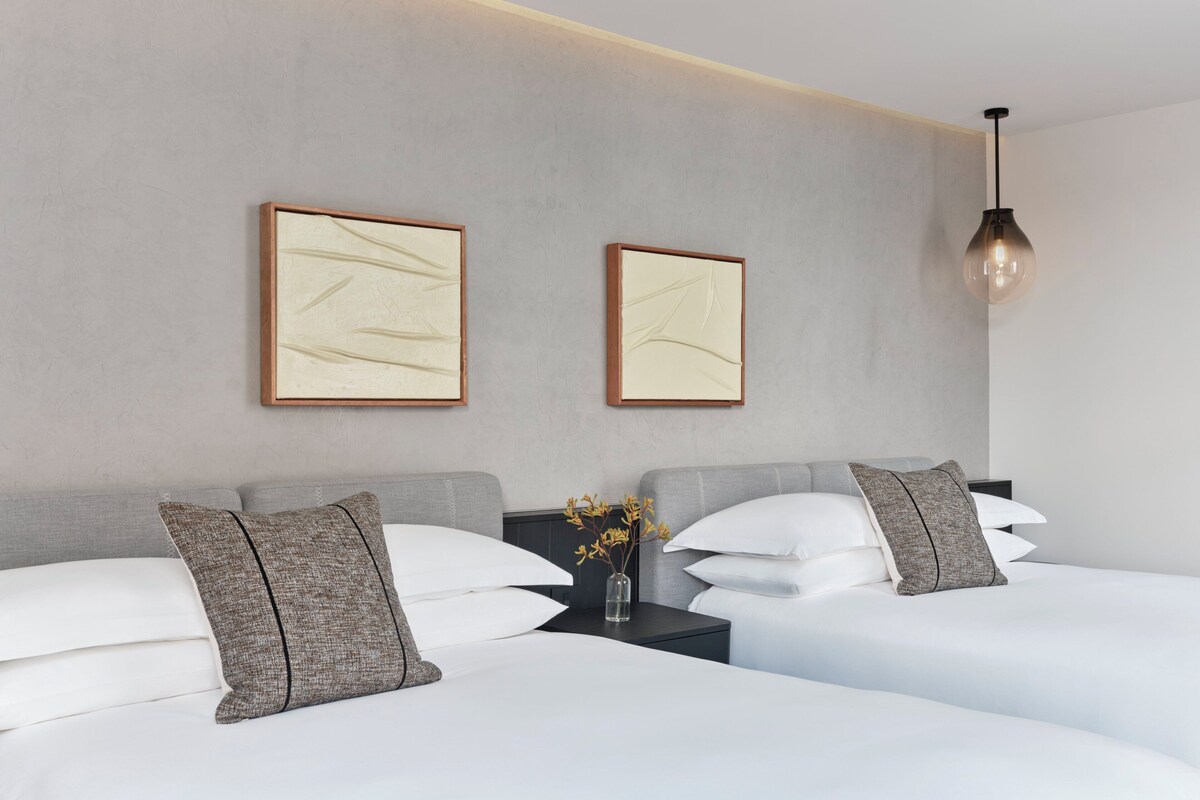 A modern hotel room with two beds, a nightstand, a vase with flowers, and two framed artworks on the wall.