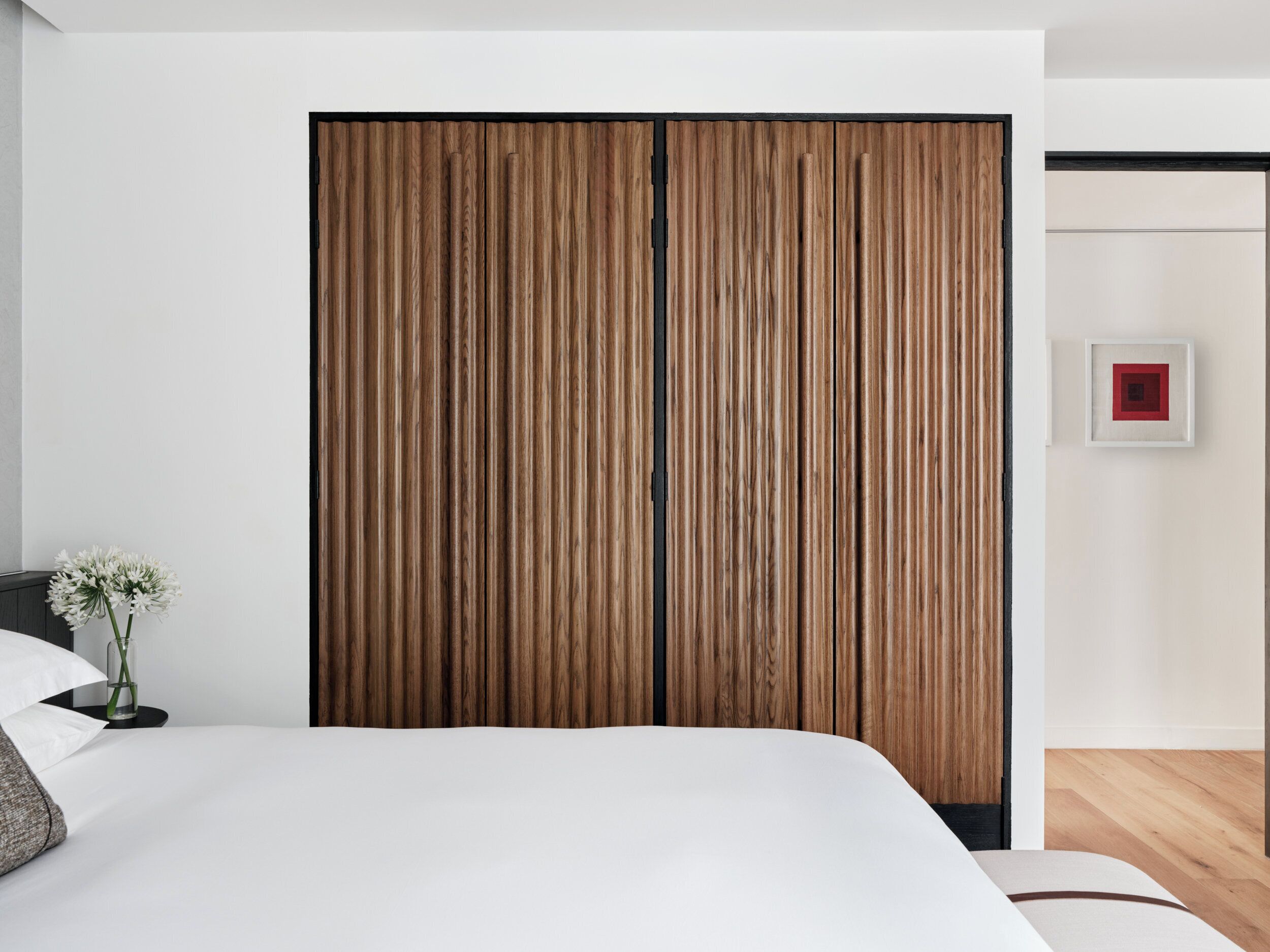 Een slaapkamer met een houten garderobe, een bed met wit beddengoed, een kleine vaas met bloemen en een ingelijst beeld aan de muur.
