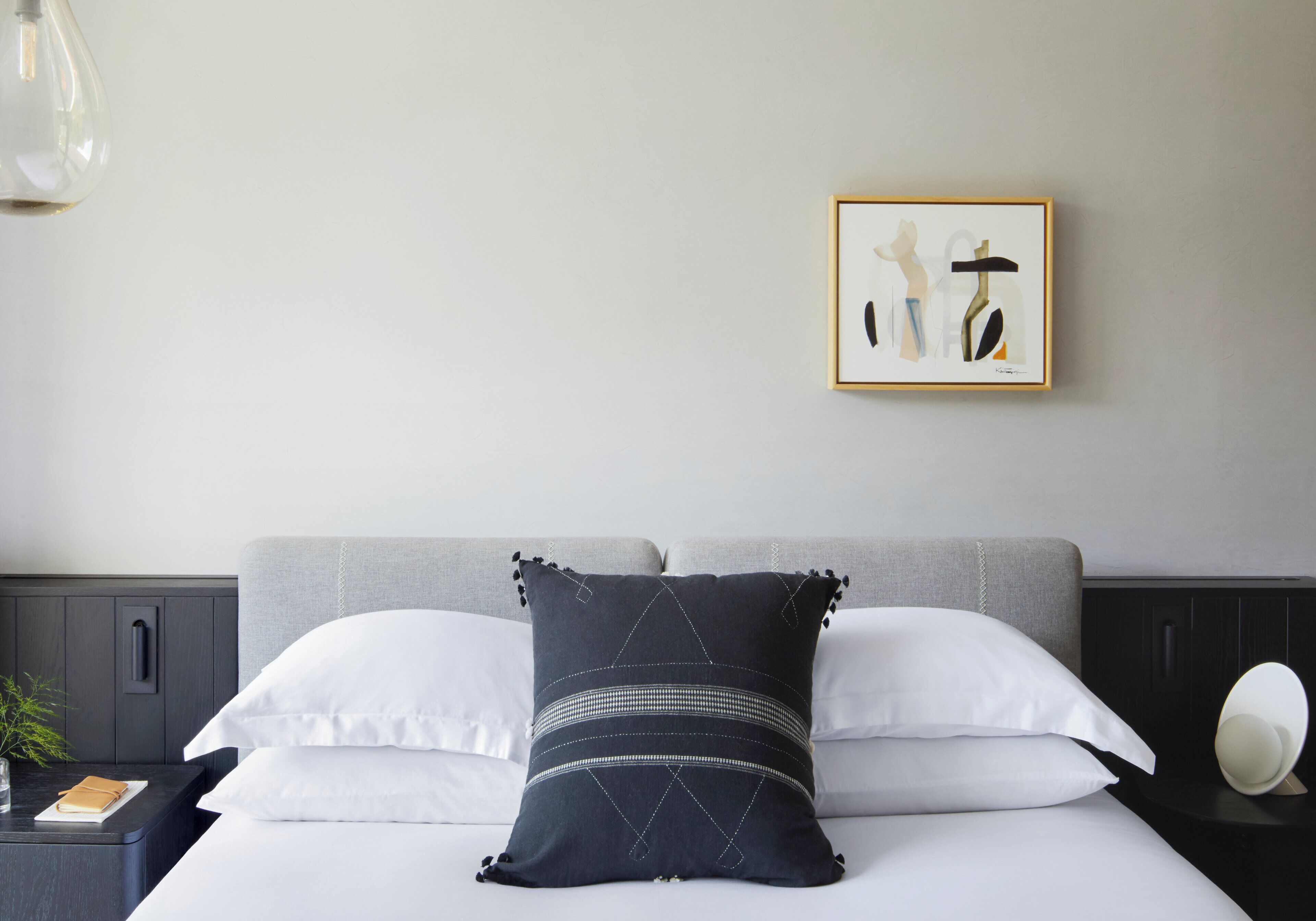 A neatly made bed with white and dark pillows, a framed artwork on the wall, and a bedside table with a lamp.