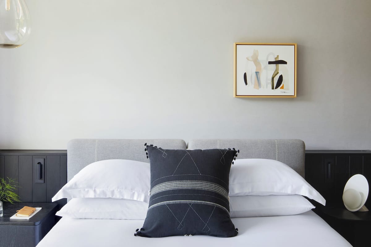 A neatly made bed with white and dark pillows, a framed artwork on the wall, and a bedside table with a lamp.