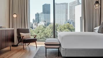 Una habitación de hotel moderna con una cama grande, una silla, una mesita y vistas de la ciudad a través de ventanales del suelo al techo.
