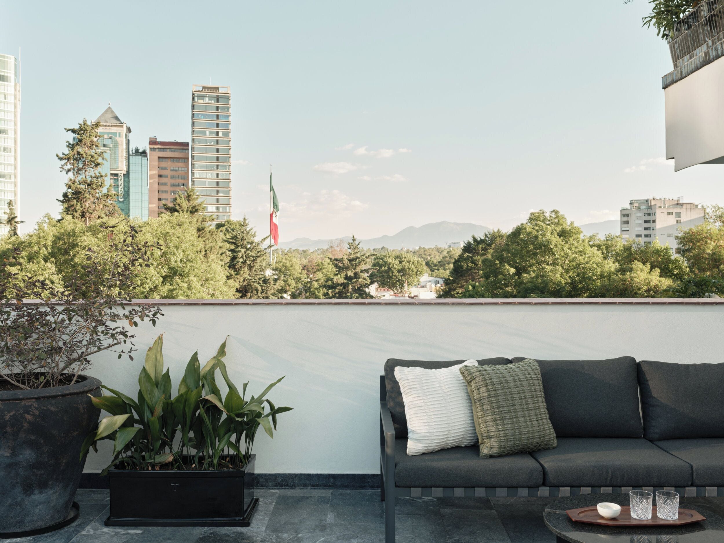 A rooftop terrace with a sofa, potted plants, and a view of city buildings and mountains.
