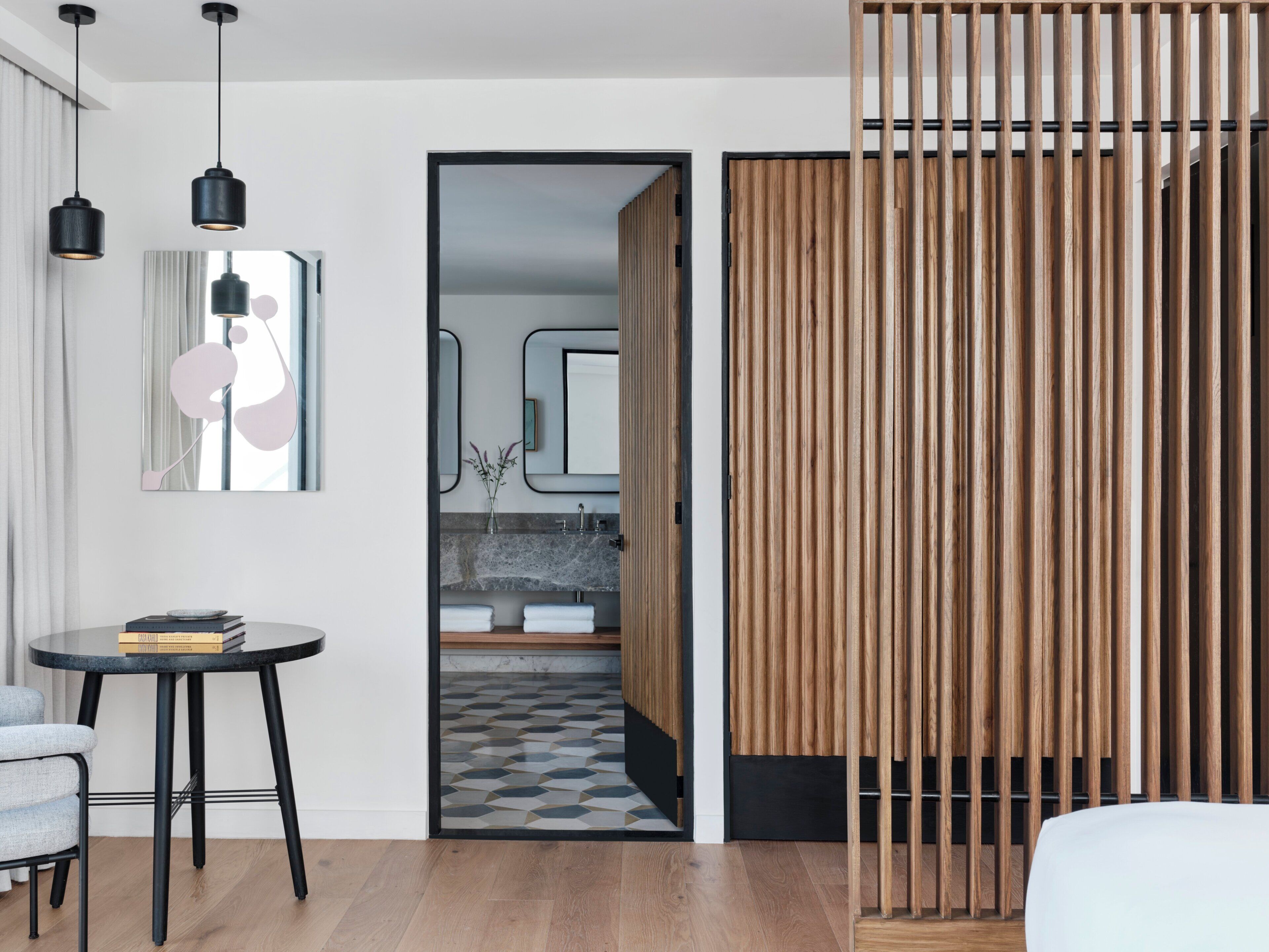 Une chambre d’hôtel moderne avec une cloison en bois, une table ronde avec des livres, un miroir et une vue sur la salle de bain.