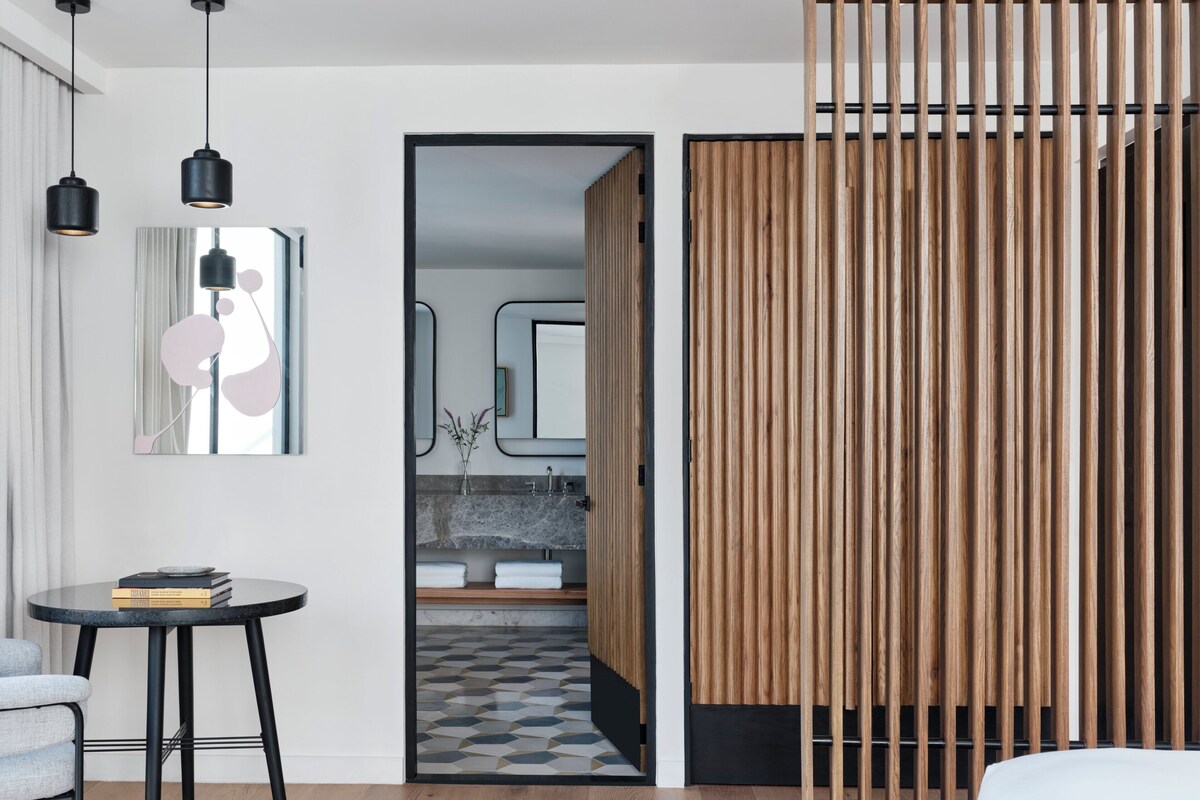 A modern hotel room with a wooden partition, a round table with books, a mirror, and a view into the bathroom.