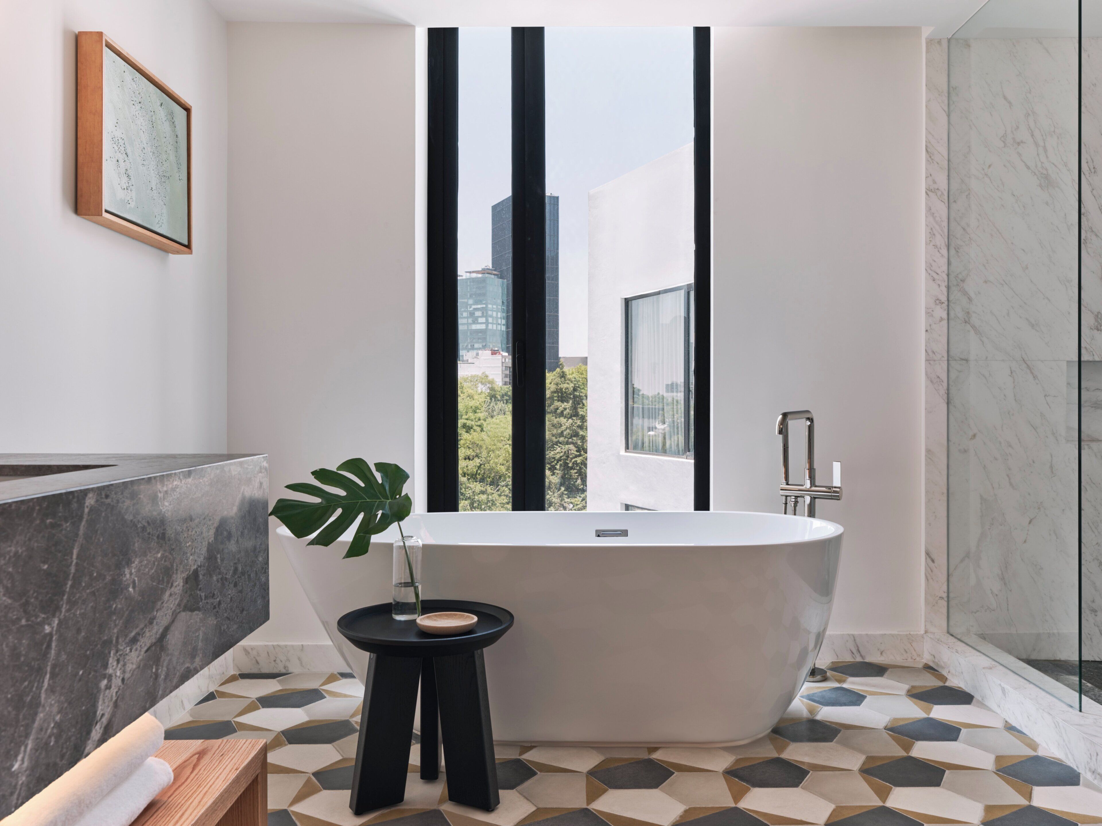 A modern bathroom with a freestanding bathtub, a small side table with a plant, and a geometric tile floor.