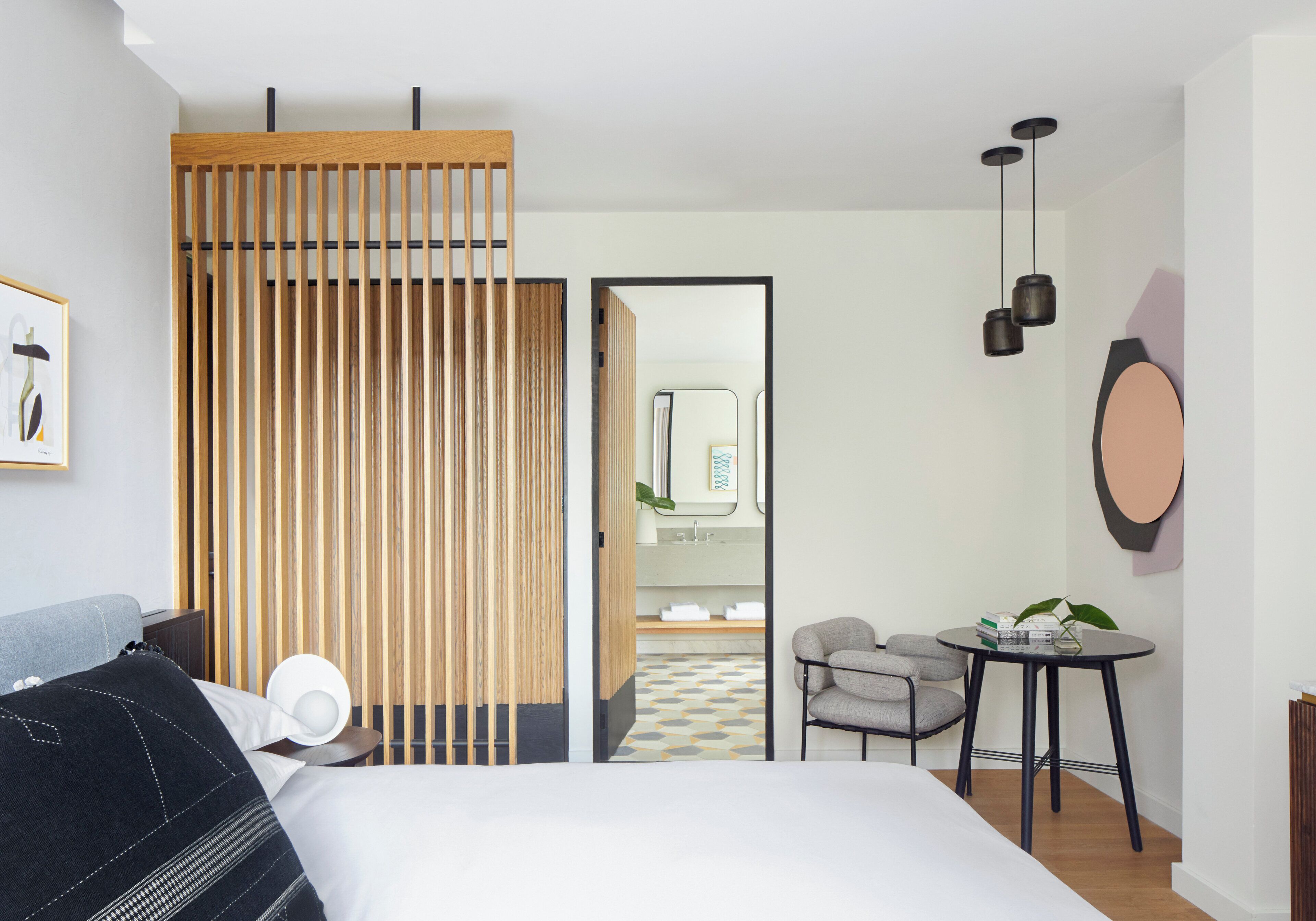 A modern hotel room with a bed, a desk, a chair, a small table, and a bathroom visible through an open door.