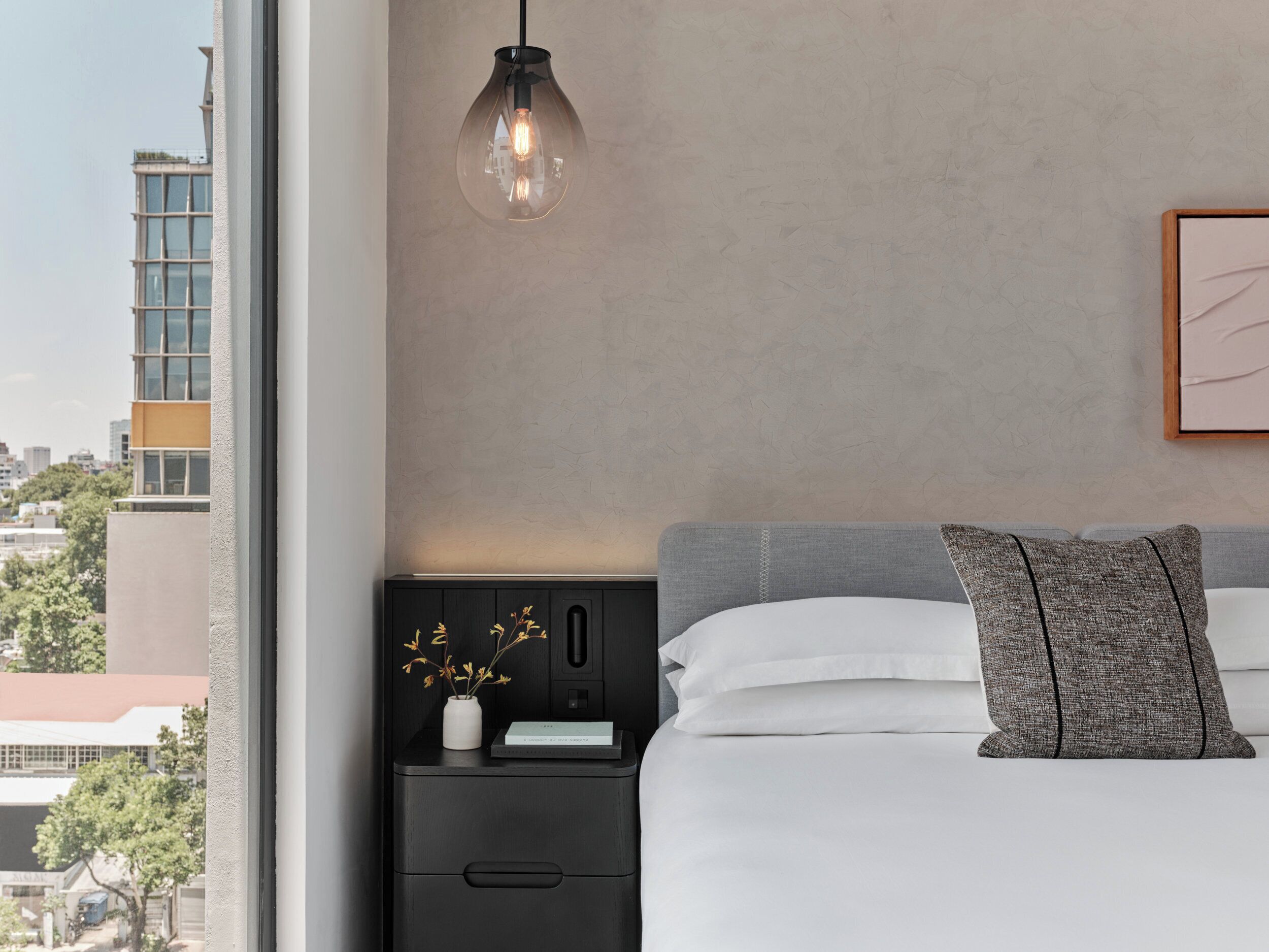 Una camera da letto moderna con un letto, un comodino, un vaso con fiori secchi, un quadro appeso alla parete e vista sulla città attraverso una finestra.