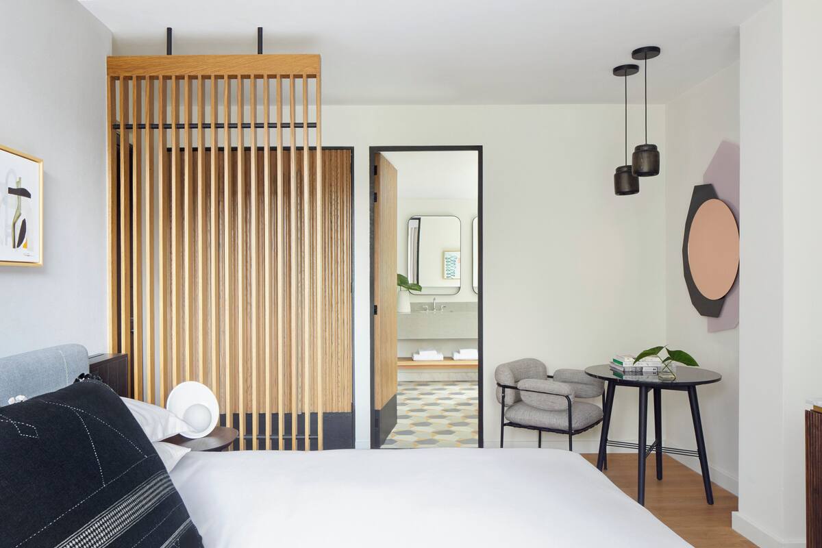 A modern hotel room with a bed, a desk, a chair, a small table, and a bathroom visible through an open door.