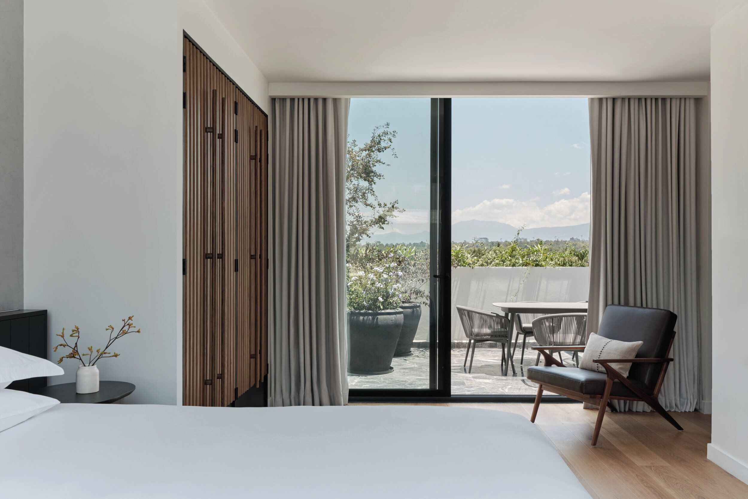 A bedroom with a bed, a chair, a table, and a sliding glass door leading to an outdoor patio with a view of greenery and mountains.