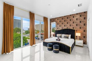 A modern bedroom with a large bed, a view of the city, and a brick-patterned wall. - Beachfront Paradise: Townhome w/ Pool and Rooftop (Pompano Beach)