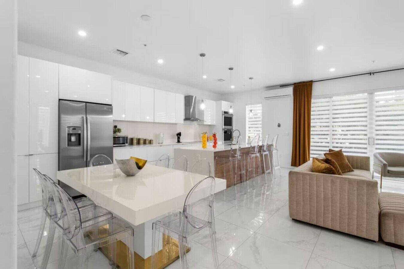 A modern kitchen with white cabinetry, a stainless steel refrigerator, and a dining area with a white table and clear chairs.