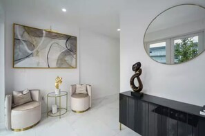 Ein modernes Wohnzimmer mit einem großen abstrakten Gemälde, zwei runden Sesseln, einem kleinen Tisch mit einer Skulptur, einer dunklen Konsole und einem runden Spiegel, der den Blick aus dem Fenster reflektiert.