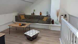 Ein Loft-Wohnzimmer mit hoher Decke, einem grauen Ecksofa, einem Holztisch, einem gemusterten Pouf und einem Dachfenster.