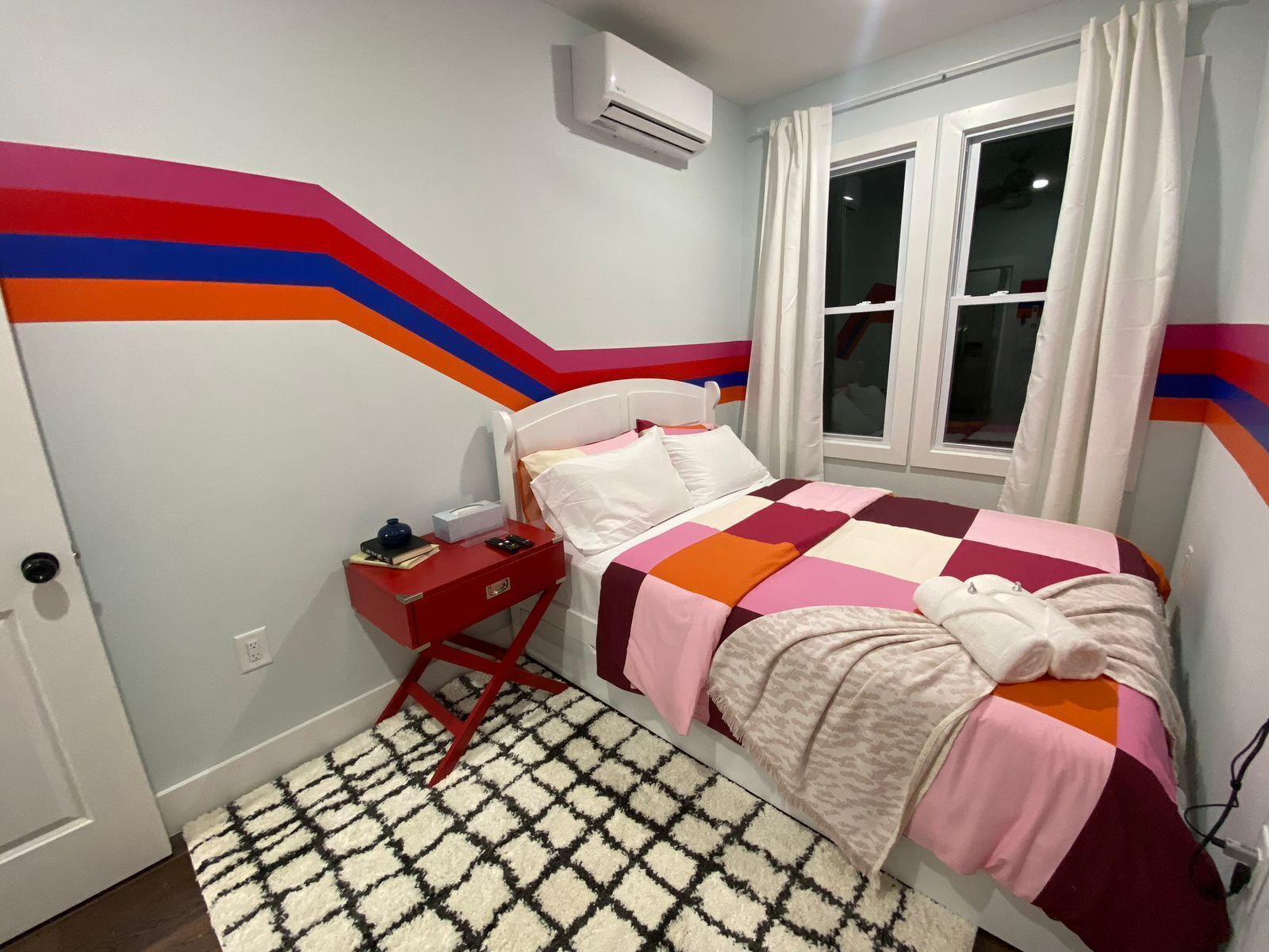 Ein Schlafzimmer mit einem Bett, einem Nachttisch, einem Fenster mit Vorhängen und einer Wand mit farbigem Design.