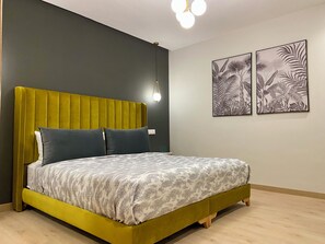 Ein Bett mit einem grünen, gepolsterten Kopfteil, Bettwäsche in Grau und Weiß mit Muster sowie zwei an der Wand befestigte Kunstwerke mit Blattmotiven.