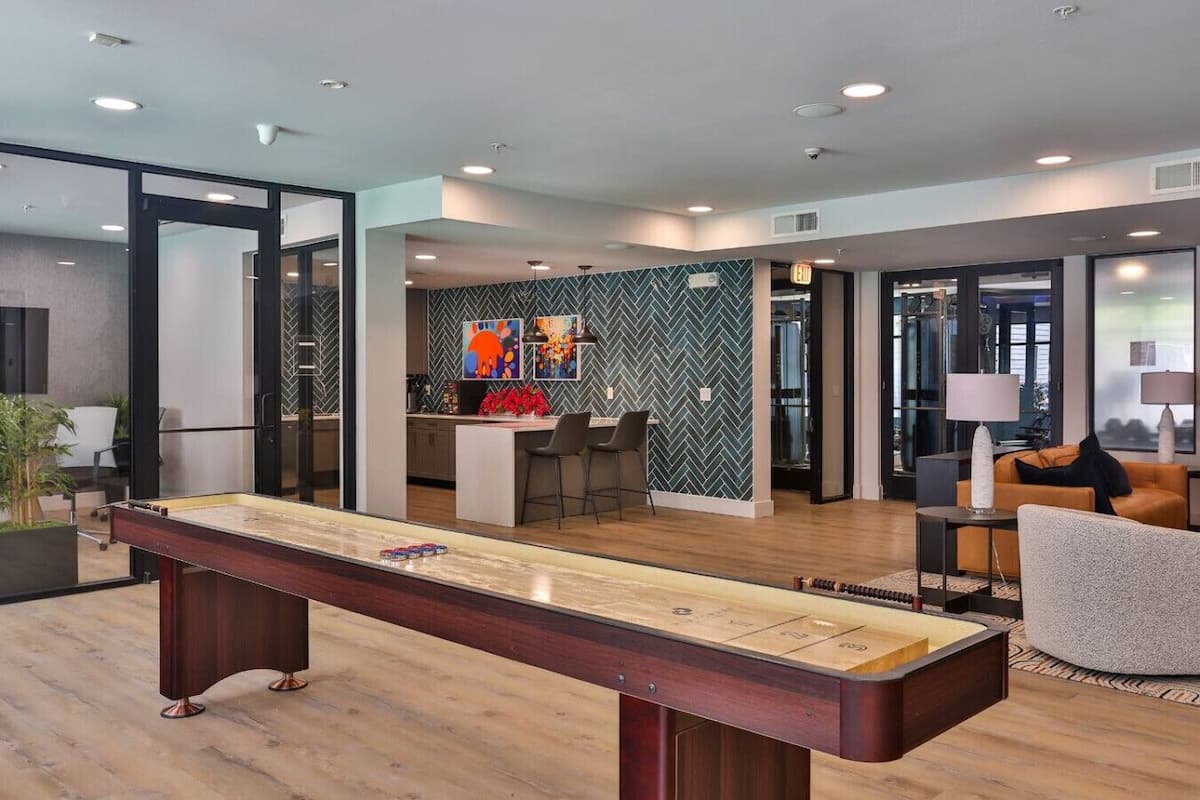 A modern living room with a shuffleboard table, a kitchen area with a herringbone-patterned backsplash, and a seating area with a sofa and chairs.
