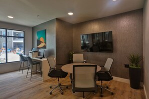 Een moderne kantoorruimte met een ronde tafel, vier stoelen, een bureau met een computer en twee grote wandmontageschermen.