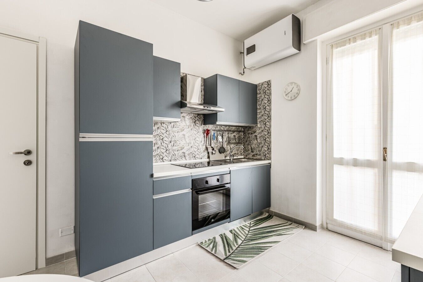 Une cuisine compacte avec des meubles gris, un plan de travail blanc, un pare-feu décoré, un four encastré et une grande fenêtre agrémentée de rideaux légers.