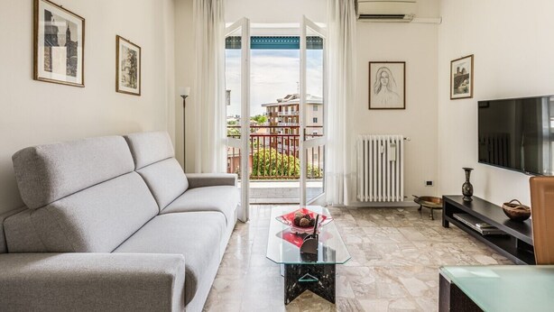A living room with a sofa, a TV, a glass coffee table, and a view of a balcony with buildings. - Maristella Holiday House (Pregnana Milanese)