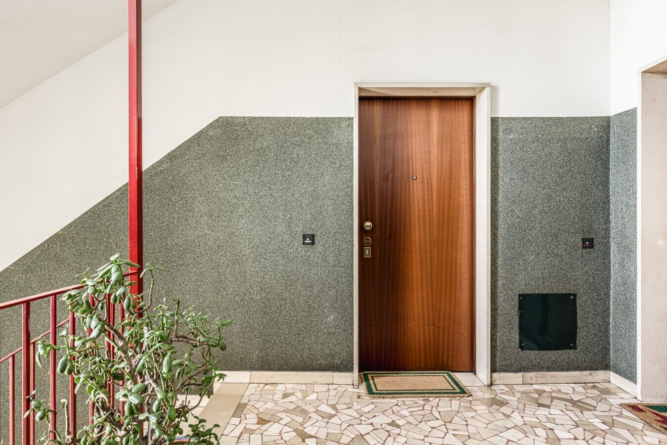 A hotel entrance with a wooden door, a red railing, a tiled floor, and a green plant.