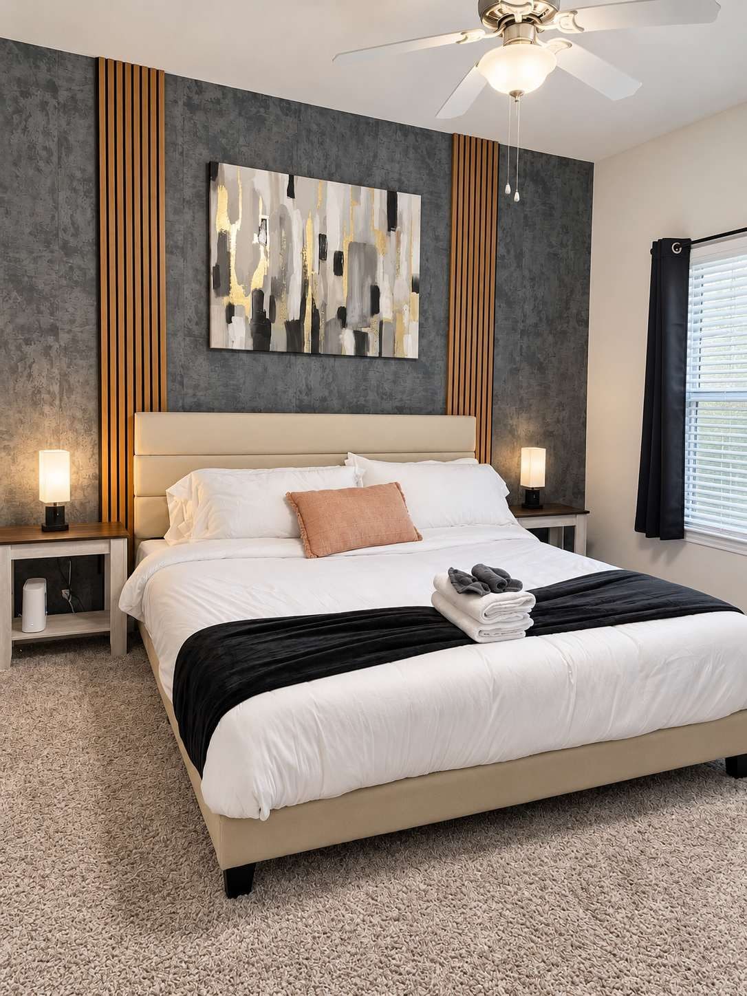 Ein modernes Schlafzimmer mit einem großen Bett, zwei Nachttischen mit Lampen, einem Deckenventilator und abstrakter Wandkunst.