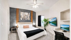 Une chambre à coucher comprenant un lit, un bureau, deux écrans d’ordinateur, un ventilateur de plafond et un grand tableau abstrait.