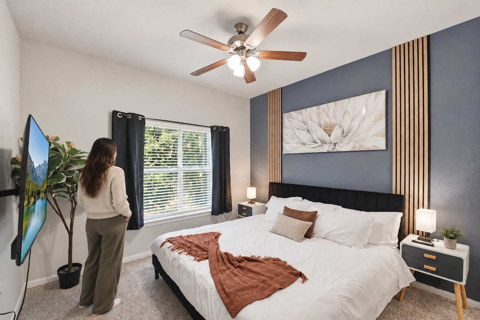 A bedroom with a bed, a ceiling fan, a window with curtains, a wall-mounted artwork, and a bedside table with a lamp.