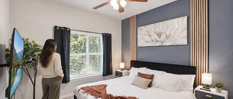 A bedroom with a bed, a ceiling fan, a window with curtains, a wall-mounted artwork, and a bedside table with a lamp.