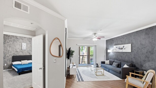 Ein modernes Wohnzimmer mit grauem Sofa, einem Holzstuhl, einem weißen Schreibtisch und einem blauen Bett im angrenzenden Zimmer.