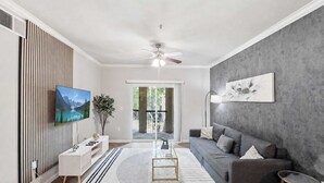Ein modernes Wohnzimmer mit Fernseher, grauem Sofa, Couchtisch und großem Fenster.