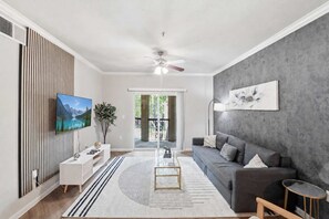 Ein modernes Wohnzimmer mit Fernseher, grauem Sofa, Couchtisch und großem Fenster.