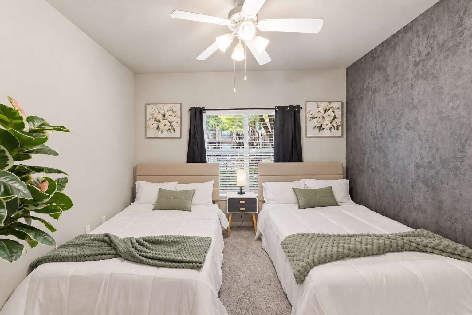 Habitación doble con dos camas, ventilador de techo, ventana con cortinas y una planta.