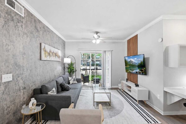 Ein modernes Wohnzimmer mit grauem Sofa, einem Glastisch, einem an der Wand befestigten Fernseher und Blick ins Freie durch große Fenster.