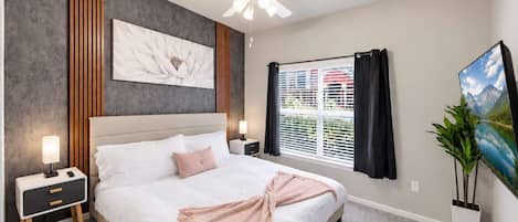 Un dormitorio con cama, mesitas de noche, ventilador de techo, ventana con cortinas y un cuadro en la pared.