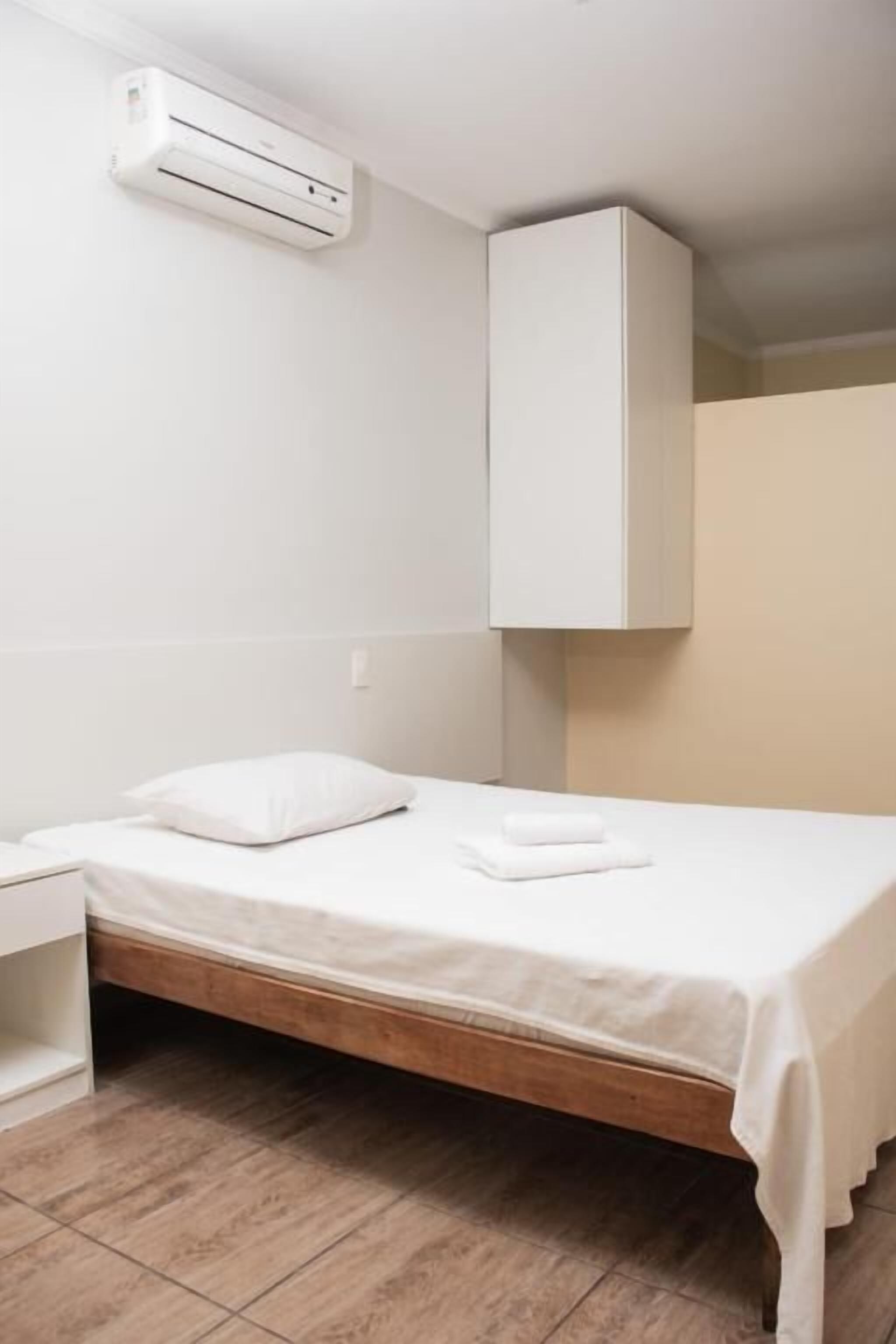 A single bed with a wooden frame, a white mattress, and a white pillow. There is a white nightstand to the left of the bed. An air conditioning unit is mounted on the wall above the bed.