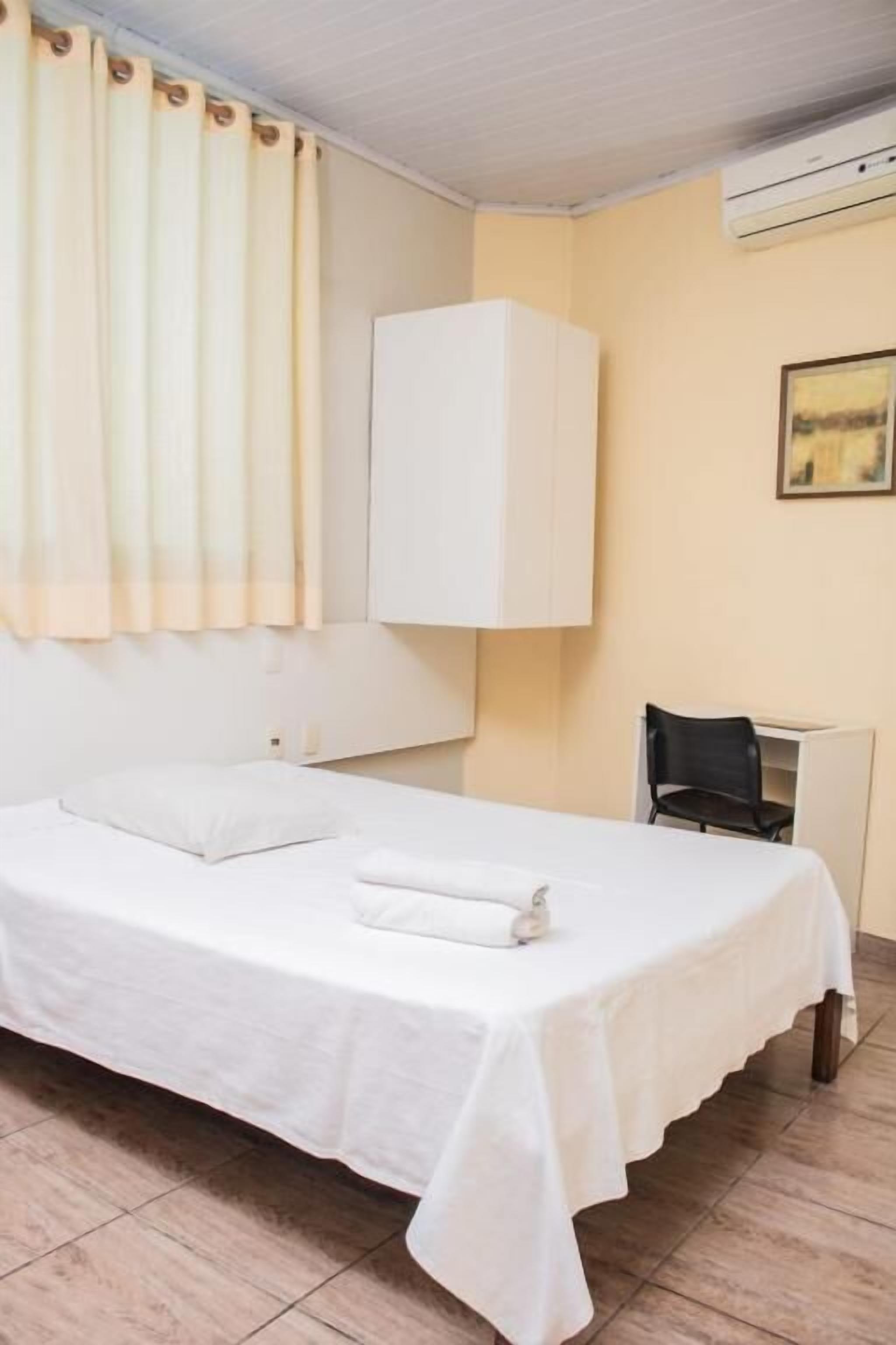 A hotel room with a bed, a white cabinet, a chair, and a wall-mounted air conditioner.