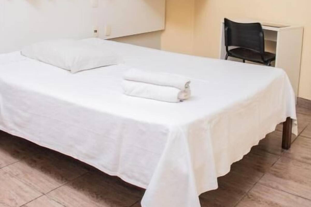 A hotel room with a bed, a white cabinet, a chair, and a wall-mounted air conditioner.