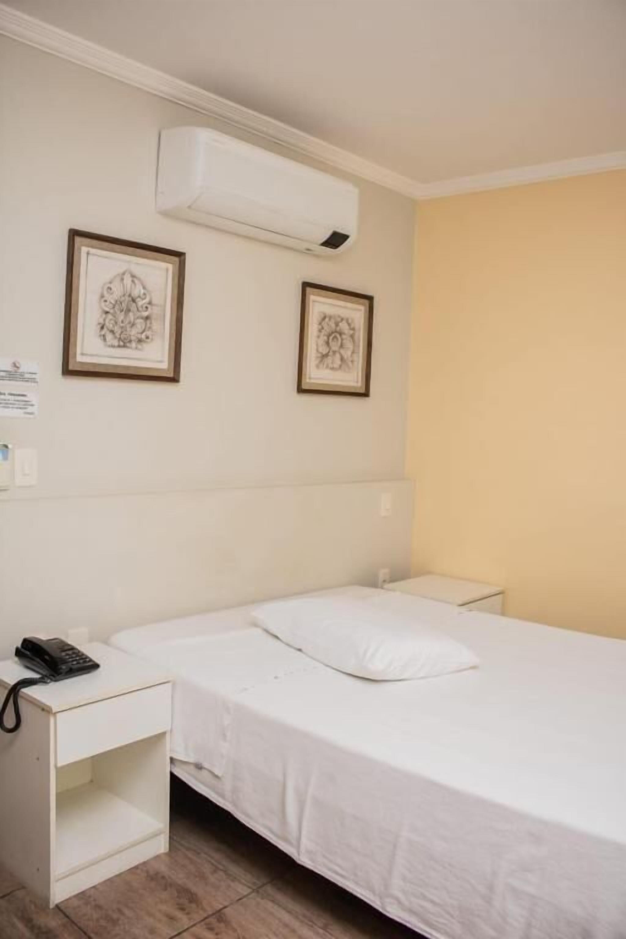 A single bed with a white mattress and pillow, a bedside table with a phone, and two framed pictures on the wall.