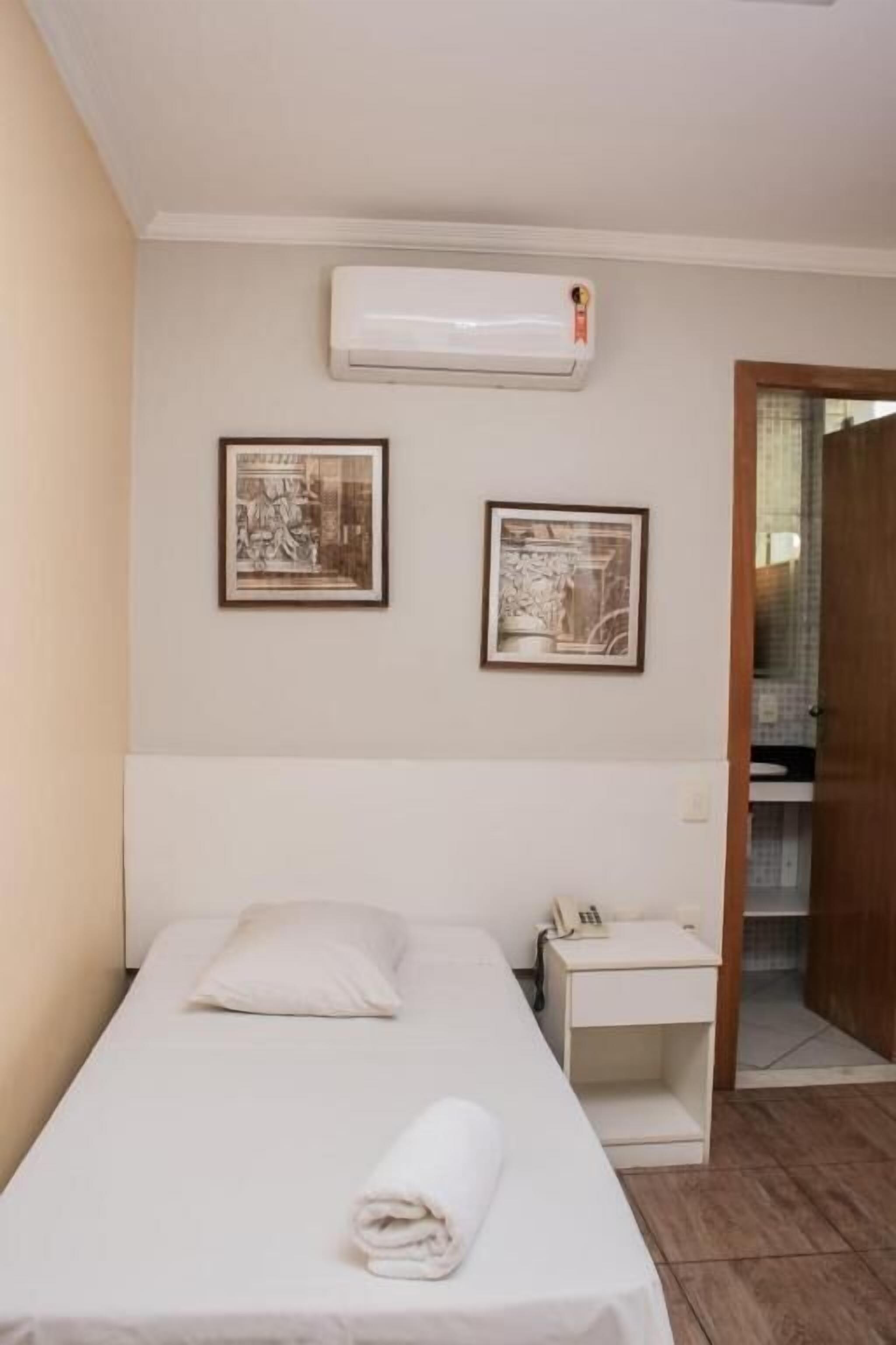 A single bed with a rolled towel, a bedside table with a phone, and two framed pictures on the wall.