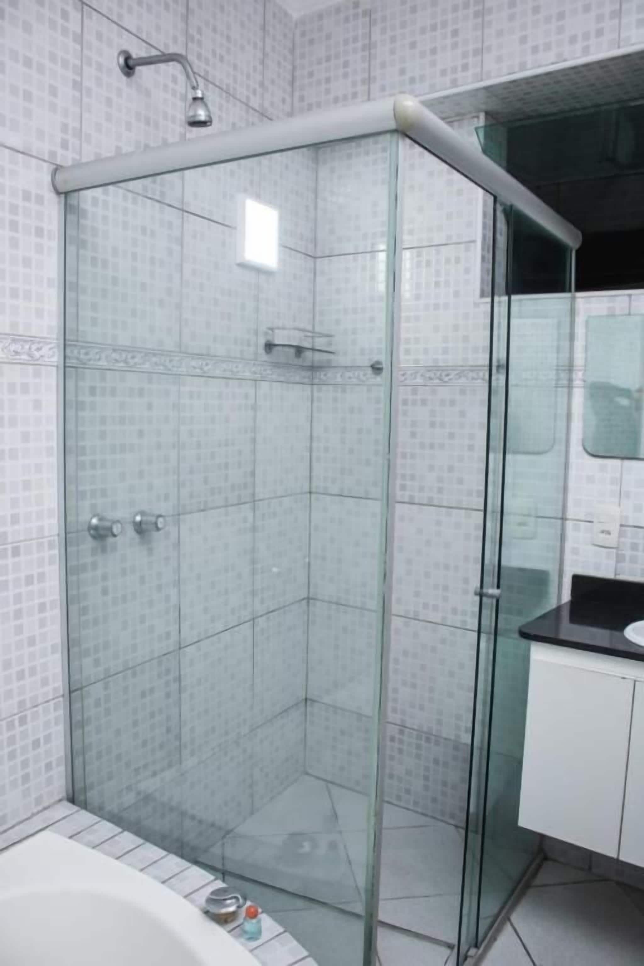 A glass-enclosed shower with a white tiled wall, a white bathtub, and a white cabinet.