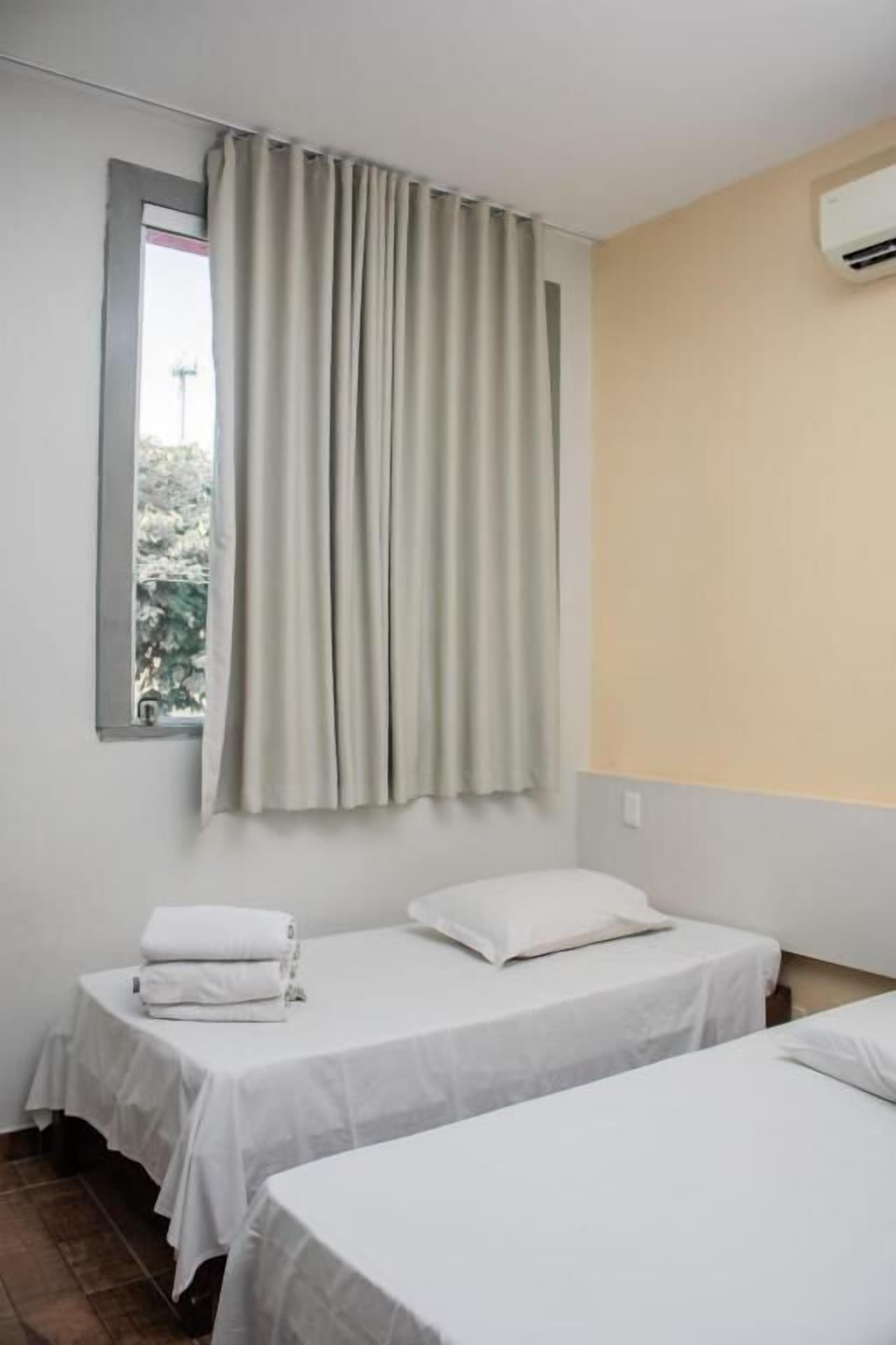 A room with two beds, a window with curtains, a wall-mounted air conditioner, and a small window with a view of greenery.