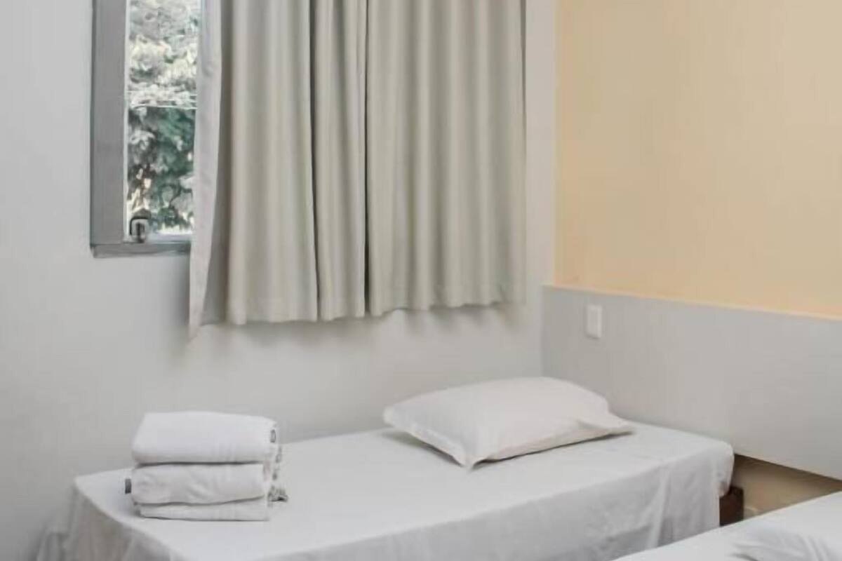 A room with two beds, a window with curtains, a wall-mounted air conditioner, and a small window with a view of greenery.