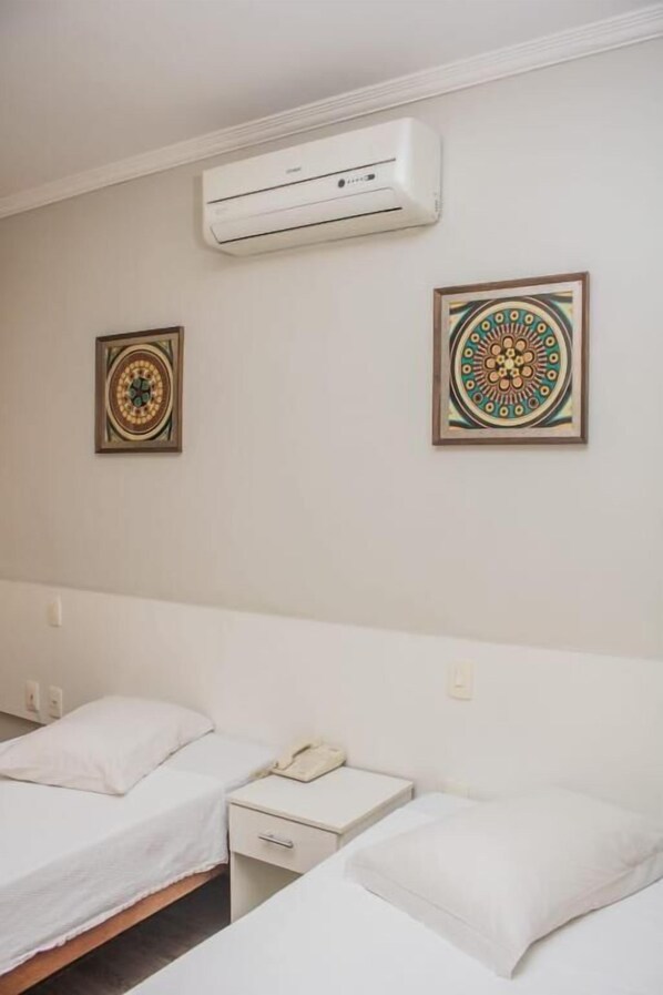 A hotel room with two beds, a bedside table with a phone, an air conditioning unit, and two decorative wall hangings.