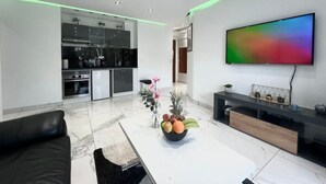 Un salon moderne avec un téléviseur à écran plat, un canapé en cuir noir, une table basse avec des fruits, et une cuisine en arrière-plan.